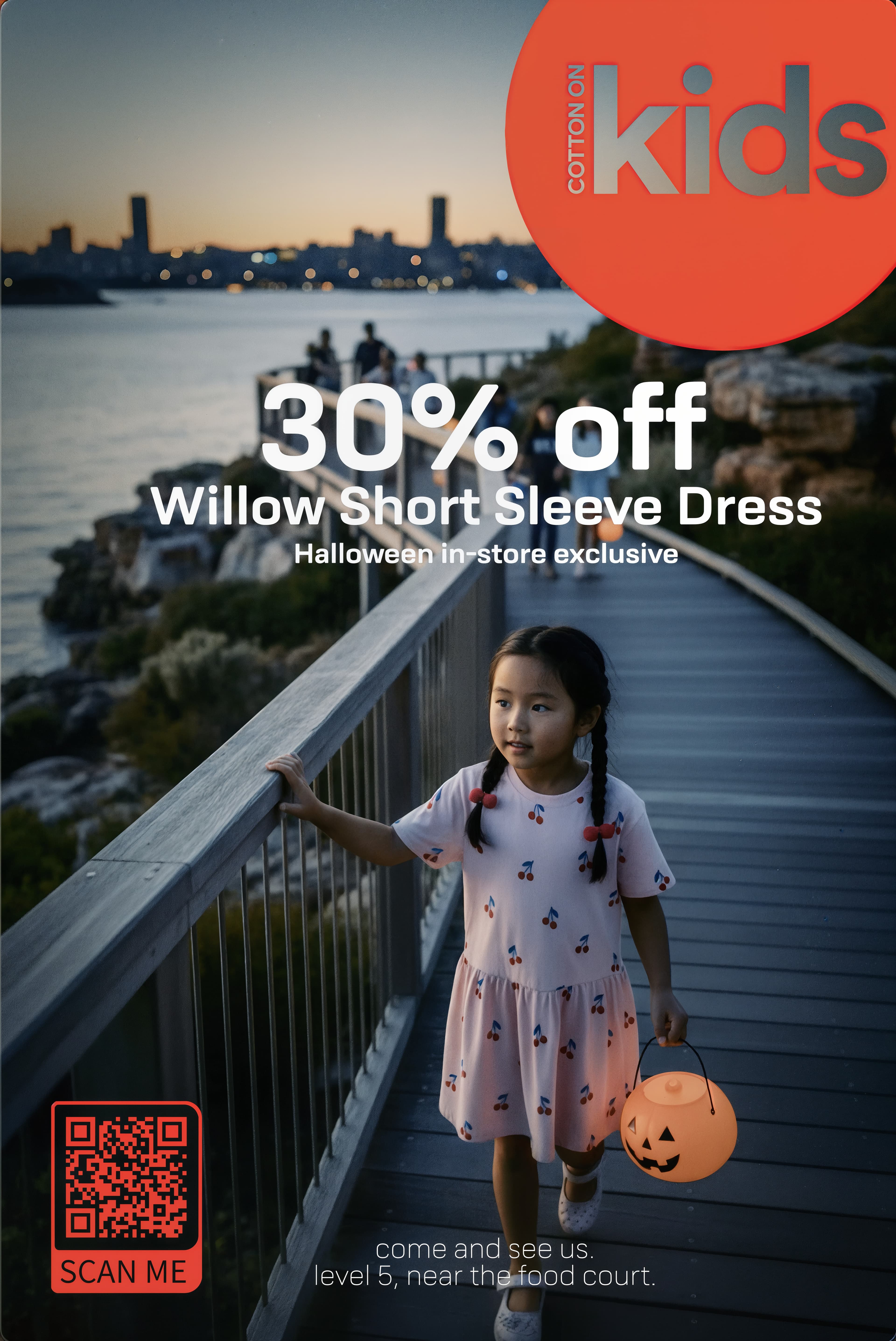 Cotton On Kids promo: girl in a cherry-print dress with a pumpkin bucket on a boardwalk at dusk